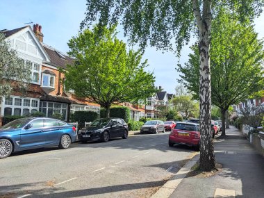 Londra, İngiltere - 04.12.2024: Yeşil ağaçları, yarı müstakil evleri ve her iki taraftaki park edilmiş arabaları olan normal bir yerleşim yeri bulutsuz mavi gökyüzü ve ilkbaharda güneş ışığı altında