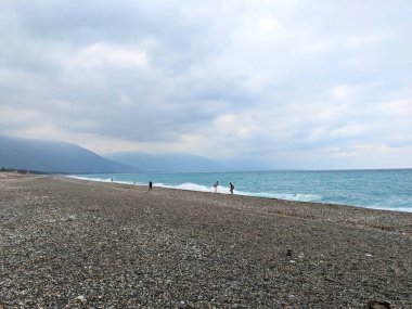 Hualien, Tayvan - 25 Mayıs 2025: İnsanlar Qixingtan 'da geniş bir çakıl taşı plajı boyunca yürürken turkuaz Pasifik Okyanusu dalgaları arka planda dağlarla birlikte dramatik bulutlu bir gökyüzü altında kıyıya çarpıyor.