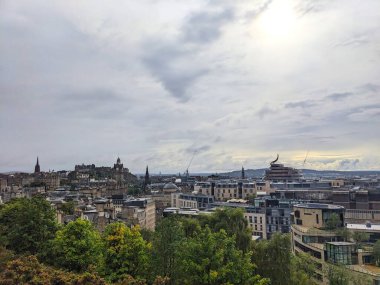 Edinburgh, İskoçya - 15 Temmuz 2025: Şehrin yüksek açılı bir görüntüsü, önplanda yeşil ağaçlarla dolu dramatik bir gökyüzü altında Eski Şehir mimarisi ile modern binalar arasındaki zıtlığı yakalıyor