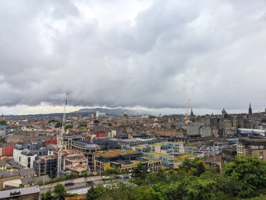 Edinburgh, İskoçya - 15 Temmuz 2025: Uzaktaki tepelerle dolu geniş, bulutlu bir gökyüzü altında tarihi mimari ve modern yapı arasındaki zıtlığı gösteren panoramik bir şehir manzarası