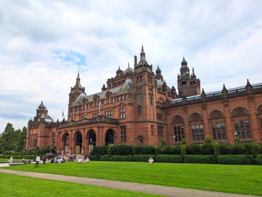 Glasgow, İskoçya - 13 Temmuz 2025: Kelvingrove Sanat Galerisi ve Müzesi 'nin çarpıcı İspanyol Barok cephesi, ziyaretçiler Kelvingrove Parkı' nda bulutlu bir gökyüzünün altında çimlerde dinleniyor