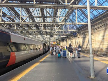 Glasgow, İngiltere - 12 Temmuz 2025: Valizli yolcular, Glasgow Central tren istasyonunda kemerli cam çatısına sahip modern şehirlerarası Avanti West Coast treninin yanındaki platformda yürüyorlar