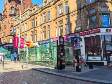 Glasgow, İskoçya, İngiltere - 12 Temmuz 2025: Yayalar Buchanan Caddesi metro istasyonunun modern cam girişinden geçerek tarihi mimarisi olan hareketli, güneşli şehir merkezi alışveriş caddesi üzerinde yürüyorlar