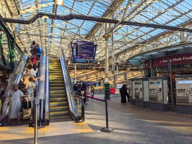 Edinburgh, İngiltere - 18 Temmuz 2025: Bavullu gezginler gün batımında geniş kemerli cam ve çelik çatı altında kalabalık bir Edinburgh Waverley İstasyonu 'nun platformlarına doğru yürüyen merdivenle gidiyorlar