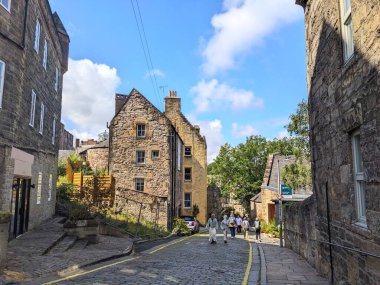 Edinburgh, İskoçya - 16 Temmuz 2025: Bir grup turist Dean Village 'da, mavi gökyüzünün altında bulutlu tarihi taş binalarla çevrili güzel bir çan kulesi boyunca yürüyorlar