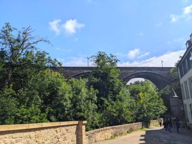 Edinburgh, İskoçya - 16 Temmuz 2025: İnsanlar tarihi Dean Village 'da taştan bir patika boyunca yürüyorlar, Dean Köprüsü, açık mavi gökyüzü altında yemyeşil yaz yapraklarıyla çevrili vadiyi kaplıyor.