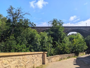 Edinburgh, İskoçya - 16 Temmuz 2025: İnsanlar, bereketli yaz yapraklarıyla çevrili Dean Köprüsü 'nün büyük kemerlerinin altında Leith' in Suyu 'nun yanında taş bir patika boyunca yürürler.