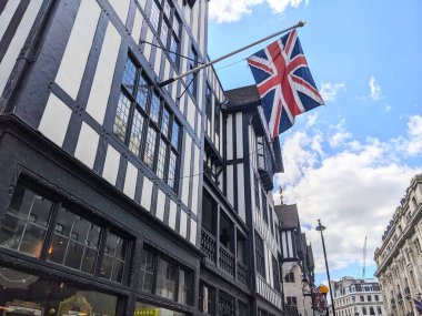 Londra, İngiltere - 09 Ağustos 2025: Batı Yakası 'nda parçalı bulutlu mavi bir gökyüzüne karşı dalgalanan Union Jack bayrağıyla Liberty mağazasının ikonik Tudor Diriliş cephesinin düşük açılı görüntüsü