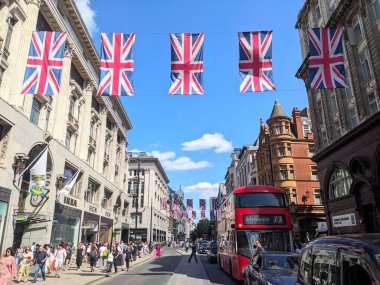 Londra, İngiltere - 09 Ağustos 2025: Klasik bir çift katlı çift katlı çift katlı otobüs, vatansever Union Jack bayraklarıyla süslenmiş kalabalık bir Oxford caddesi boyunca mavi gökyüzü altında yürüyen bir kalabalık gibi yürüyor.