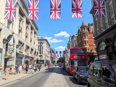 Londra, İngiltere - 09 Ağustos 2025: Oxford Caddesi 'nde canlı bir şehir manzarası, kaldırımda müşteriler ve trafikte kırmızı bir otobüs, açık bir yaz gününde mavi gökyüzü altında sayısız İngiliz bayrağı altında