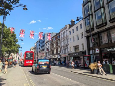 Londra, İngiltere - 09 Ağustos 2025: Klasik bir kırmızı çift katlı otobüs ve hareketli Oxford Caddesi boyunca siyah bir taksi, mavi gökyüzünün altında Union Jack bayraklarıyla süslenmiş ünlü bir alışveriş merkezi.