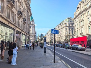 Londra, İngiltere - 09 Ağustos 2025: Müşteriler, güneşli bir yaz gününde, şehir trafiği ve açık mavi gökyüzü altında bir bisikletçiyle ünlü Regent Caddesi 'ndeki mağazaların ve klasik binaların önünden geçiyor