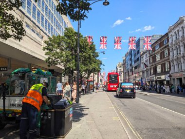 Londra, İngiltere - 09 Ağustos 2025: Bir temizlik işçisi, kırmızı çift katlı otobüs ve yayalar Union Jack bayrakları altında ünlü alışveriş caddesi boyunca ilerlerken kalabalık bir Oxford Caddesi 'ne bir çöp kutusu boşalttı