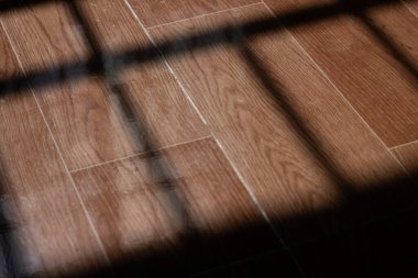 Pencere ızgarasının gölgesi güneş ışığıyla aydınlatılmış, ahşap desenli bir zemine düşer. Tutsaklığı, yalnızlığı, umudu ve zaman akışını sembolize eden çarpıcı, yüksek kontrastlı bir görüntü..