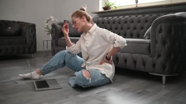 Gözlüklü sarışın bir kadın kanepenin yanında oturuyor ve evde dizüstü bilgisayarla çalışıyor. Yüksek kaliteli FullHD görüntüler