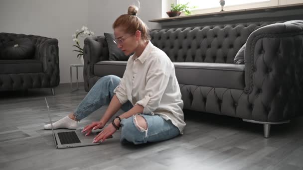 メガネのブロンドの女性はソファーの近くの床に座り ラップトップで自宅で働いています 高品質のフルHd映像 — ストック動画