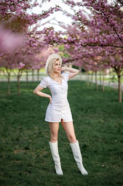 Beyaz elbiseli güzel bir sarışın çiçek açan ağaçların arkasında bir parkta duruyor. Yüksek kalite fotoğraf