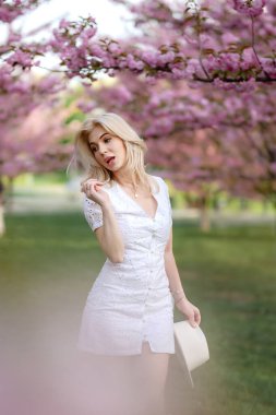 Beyaz elbiseli güzel bir sarışın çiçek açan ağaçların arkasında bir parkta duruyor. Yüksek kalite fotoğraf
