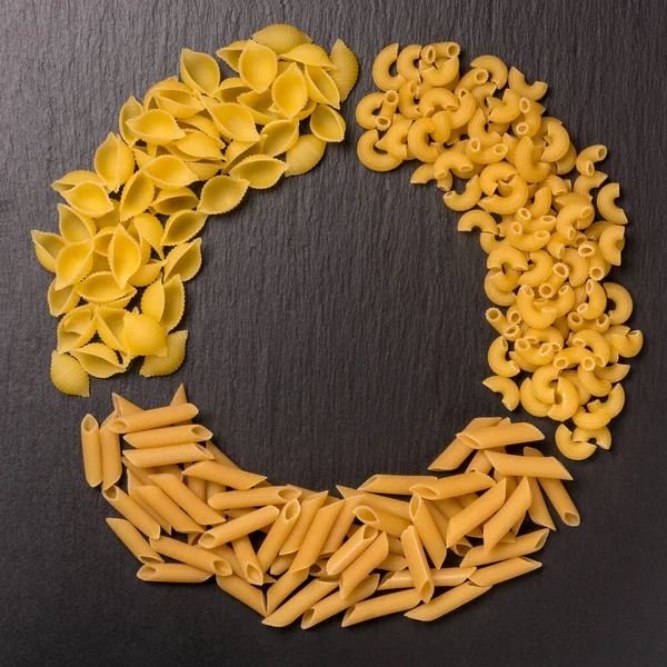 variety of uncooked pasta arranged in circle, isolated on black slate background, diet and food concept, top down view with copy space