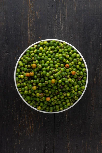 green raw black peppercorns in a white bowl, spicy and seasoning ingredient isolated on black textured background, taken from above with copy space