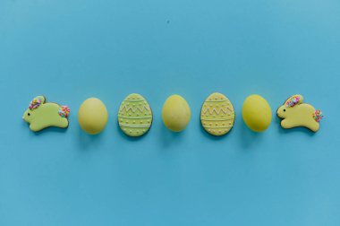 Yellow painted chicken eggs and yellow cookies in the shape of a rabbit and eggs on a blue backgroung. Top view