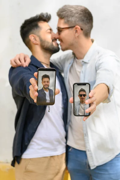 在阳光明媚的大街上 穿着休闲装的一对积极的同性恋夫妇一边微笑着一边拥抱 一边展示着带着自拍的手机 — 图库照片