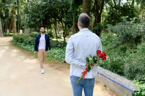 难以辨认的男人背着红玫瑰在后边等待着男人的灵魂伴侣在绿树成荫的公园里漫步 — 图库照片