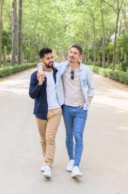 Yeşil parktaki uzun ağaçların arasında yürürken el ele tutuşup kucaklaşan, birbirine aşık, çok ırklı, genç bir çift.