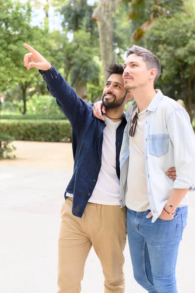 在绿色公园的阳光明媚的日子里 快乐的 留着胡子的西班牙同性恋者一边微笑一边指指点点 一边拥抱男朋友 — 图库照片
