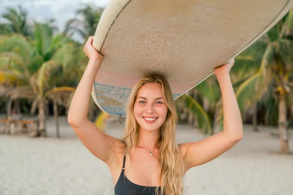 Stock image Happy cheerful young woman with long blond hair in black bikini carrying surfboard on her head at sunsets and looking at camera