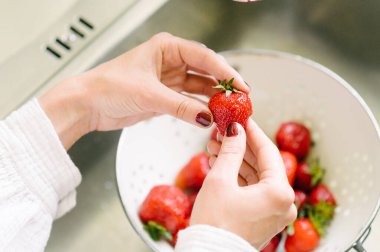Elinde taze kırmızı çilek olan kimliği belirsiz bir kadın mutfakta kahvaltıya sağlıklı bir şeyler hazırlıyor.