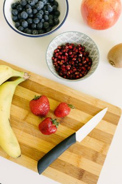 Yukardan taze olgun muzlar, çilek ve yaban mersinli nar ve kivi karışımı. Masanın üzerinde meyve suyu için tahta ve bıçak var.