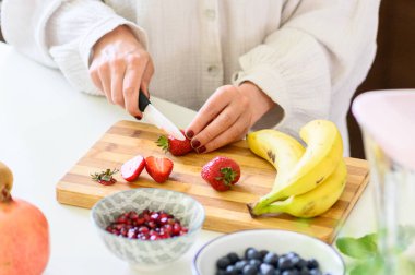 Beyaz gömlekli meçhul kadın olgun çilek ve meyve keserken mutfak tezgahında taze muzlarla sağlıklı bir kahvaltı hazırlıyor.
