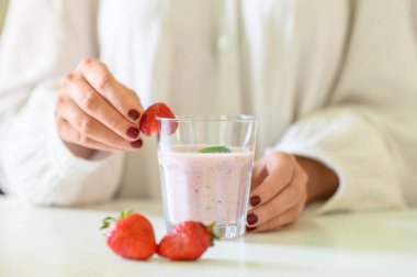 Lezzetli meyveli smoothie ile isimsiz bir kadın masada duruyor ve bir dilim çileği mutfağın kenarına koyuyor.