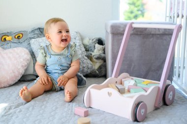 Oyuncak arabasının yanında oturan, yatak odasındaki rahat yatağın üzerinde oturan ve gözlerini kaçıran gündelik kıyafetlerle dolu neşeli küçük bir bebek.