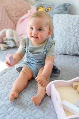 Evde rahat bir yatakta oturan, günlük giysiler içinde oyuncak bloğu olan küçük şirin bir bebek.