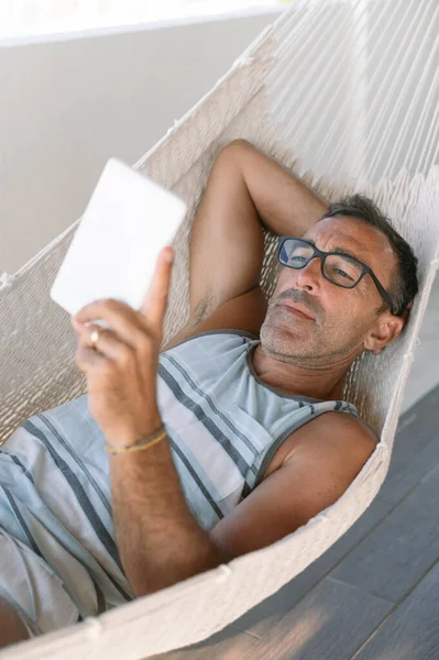 Modern ambiyansı olan şık bir çatı terasında 50 yaşında bir adam rahat bir hamak kurmuş. Elinde e-kitap okuyucusuyla. Yüksek kalite fotoğraf, tatil konsepti.