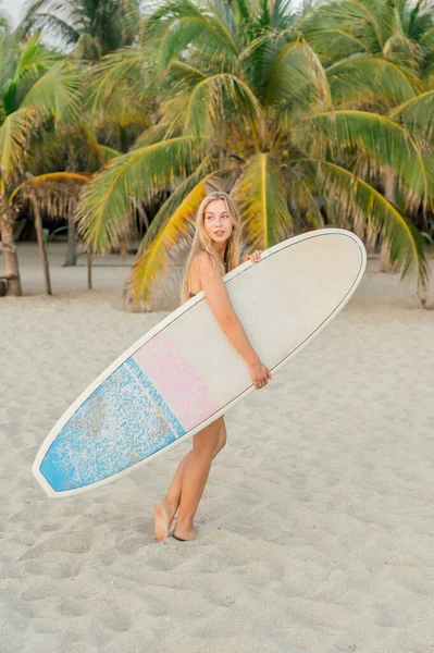 背の高い若いです素足の女性とともに長いブロンドの髪とサーフボード離れて見ている間に立って砂のビーチに対してヤシ — ストック写真