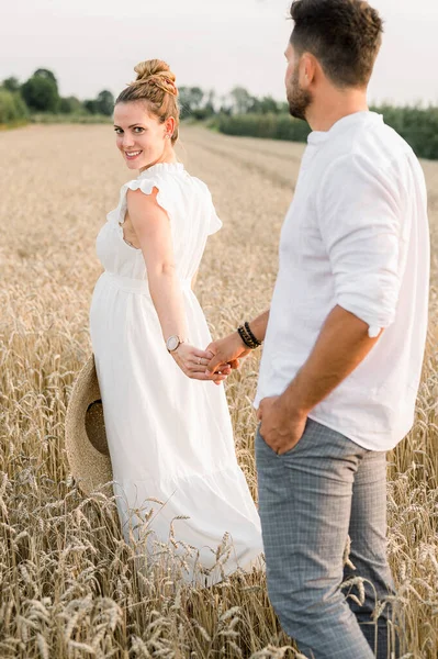 白いドレス姿の若い妊婦の側面図は 穀物畑を歩いている間に夫の手を取り合い お互いを見る — ストック写真