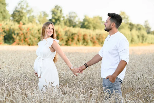 年轻的丈夫和穿着白衣的怀孕妻子手牵手站在青草丛生的草地上相互凝视的侧影 — 图库照片