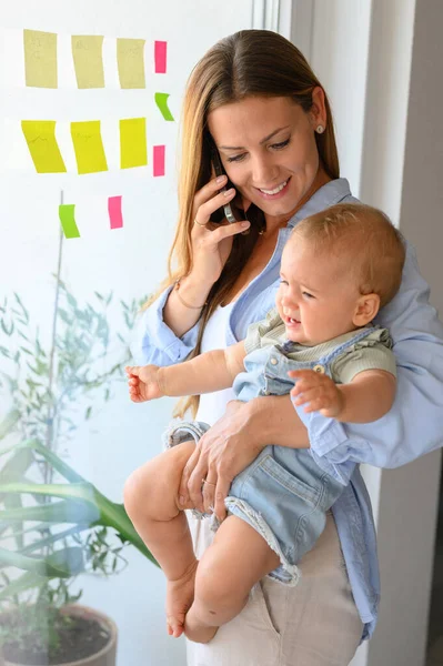 Lykkelig Ung Mor Søt Smårolling Hverdagsklær Smilende Telefonsamtale Mot Vindu – stockfoto