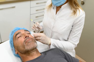 Üniformalı isimsiz kadın kozmetik uzmanı estetik kliniğinde yatakta yatan erkek hastaya yüz gerdirme ameliyatı yapıyor.