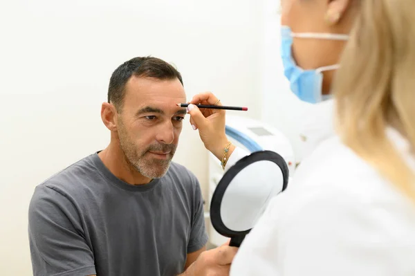 Kozmetik kliniğindeki yansımaya bakarken elinde aynayla oturan yetişkin bir erkek hastanın yüzünde kalem izi olan maskeli genç bir kozmetik uzmanı.