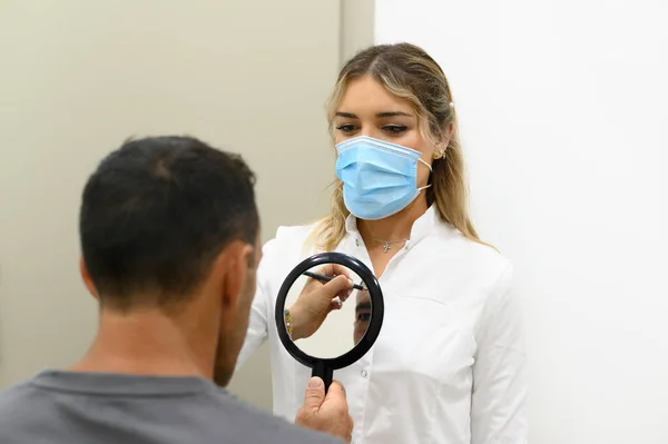Maskeli genç bir kadın kozmetik uzmanı elinde aynayla kozmetik kliniğine bakarken tanınmayan bir erkek hastanın yüzüne kalem izi bırakıyor.