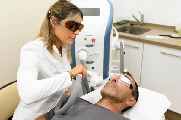 Kahverengi gözlüklü kadın kozmetolojistin modern kozmetik kliniğinde koruyucu gözlükleriyle yetişkin hasta kaşlarının fotoğrafını çekmek için lazer aletinin sapını kullanışının yan görüntüsü.