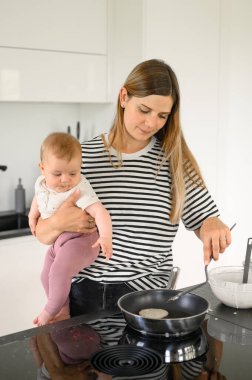 Pozitif genç anne, günlük kıyafetlerle kucağında bebek kız, fırının yanında duruyor ve mutfakta tava pişiriyor, kahvaltı için gözleme hazırlıyor. aile kavramı