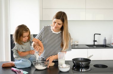 Mutfak tezgahında günlük giysiler içinde hamuru unla karıştıran, sabah kahvaltısı için evde yemek hazırlayan pozitif genç bir kadın.