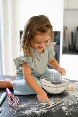Gündelik giysiler içinde küçük tatlı bir kız mutfak tezgahında oturmuş kahvaltıda krep yapmak için annesiyle hamur hazırlarken unla oynuyor.