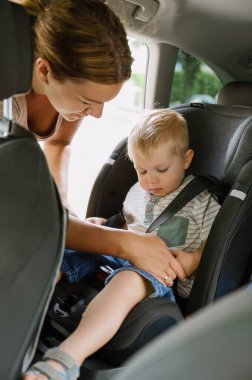 Oğlunu çocuk koltuğuna emniyet kemeriyle bağlayan günlük kıyafetli genç bir kadının yan görüntüsü. güvenlik kavramı