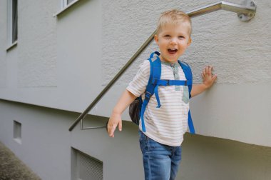 Merdivenin yanında duran ve sırt çantası takıp kameraya gülümseyen sevimli çocuğun ön görüntüsü..